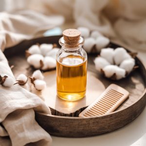 A beautifully styled flat lay featuring a glass bottle of golden castor oil on a rustic wooden tray. Surrounding the bottle are natural elements like cotton pads, a soft linen cloth, and a small wooden comb. The lighting is soft and warm, creating a cosy, spa-like atmosphere that evokes feelings of self-care and nourishment.